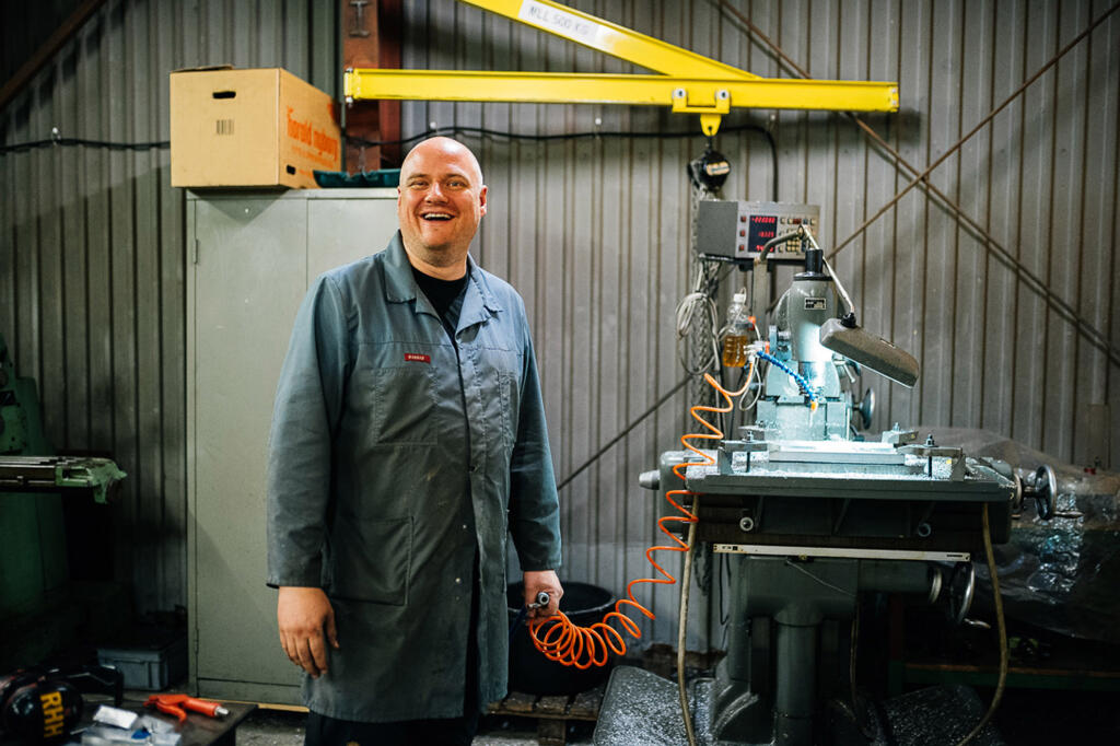 Ein Mann in grauem Arbeitskittel steht in einer Werkstatt neben einer Maschine. Er lächelt und hält ein Luftschlauch in der Hand. Im Hintergrund sind Werkzeuge und eine große Metallmaschine zu sehen.