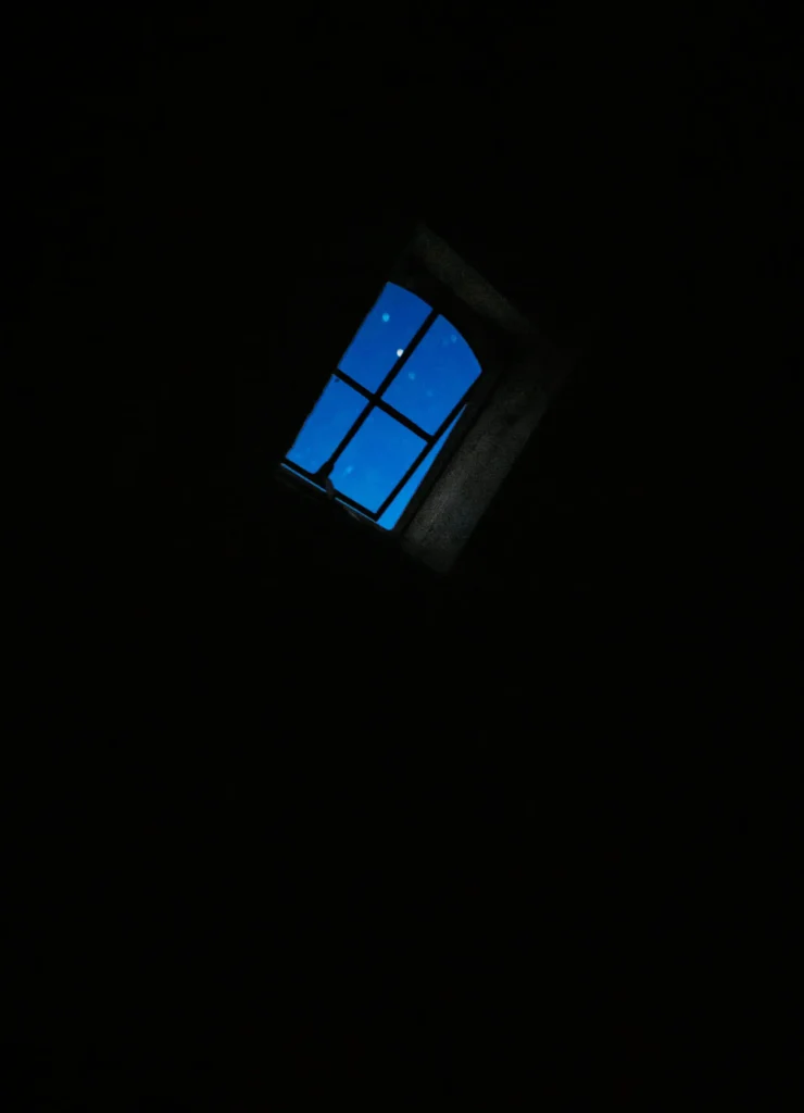 Ein Fenster in einem dunklen Raum mit Blick auf einen klaren blauen Nachthimmel und einige sichtbar funkelnde Sterne. Die Fensterrahmen sind gut erkennbar.