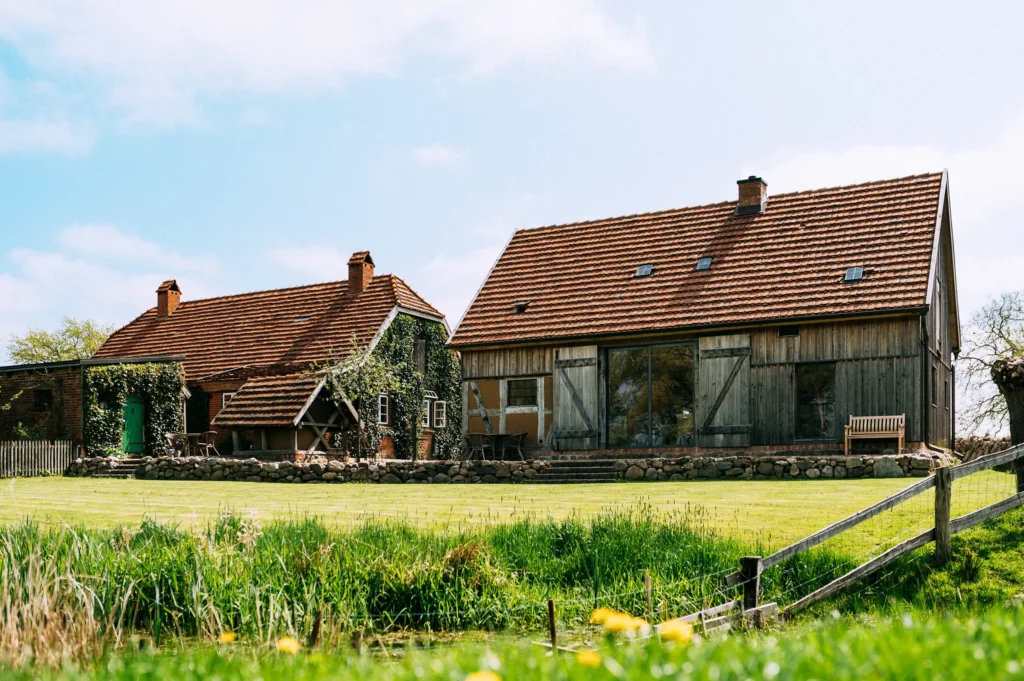 Zwei Bauernhäuser in einer grünen Landschaft mit Wiese und einem Zaun im Vordergrund. Eines der Häuser hat große Fenster, während das andere kleiner ist und mit Pflanzen bewachsen ist. Der Himmel ist blau mit einigen Wolken, was eine ruhige ländliche Atmosphäre schafft. Keywords: meine kleine farm, green, wildyard.