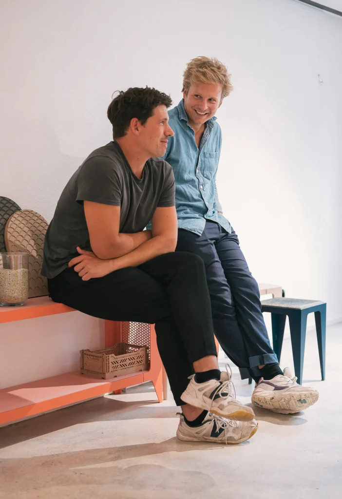 Zwei Männer sitzen auf einer Bank in einem hellen Raum. Der Mann links trägt ein graues T-Shirt und schwarze Hosen, während der Mann rechts ein blaues Hemd und dunkle Hosen anhat. Im Hintergrund sind Möbel zu sehen. Der Raum hat eine moderne, minimalistische Einrichtung.
