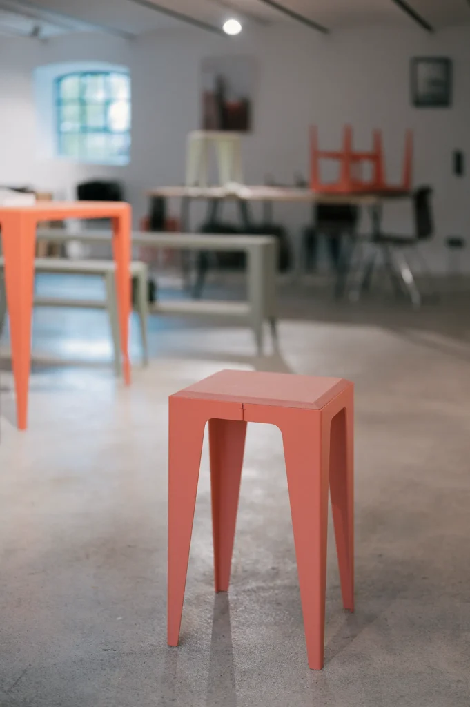 Ein oranger Hocker mit schmalen Beinen steht in einem modernen Raum. Im Hintergrund sind weitere Möbel in verschiedenen Farben wie grün und beige zu sehen. Der Raum hat einen minimalistischen Stil und wirkt hell.