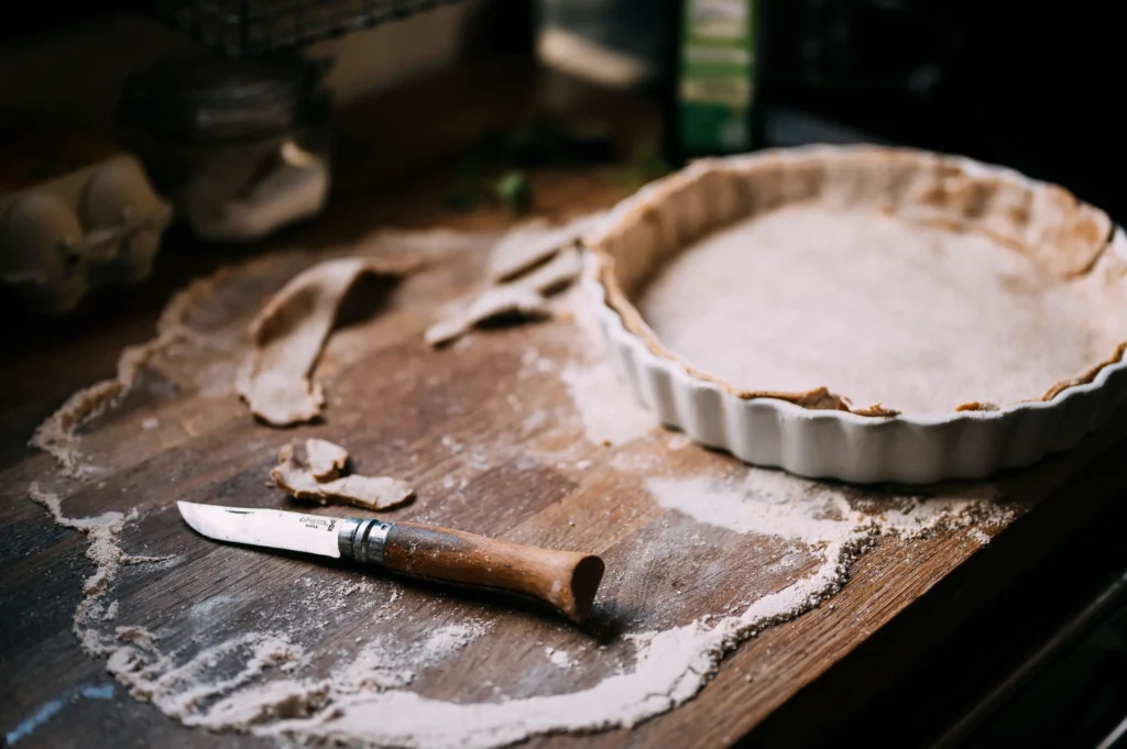 Eine Holzarbeitsfläche mit Mehl, einem Teigroller und einem bereitgestellten Tarteboden in einer weißen Form. Ein Messer mit Holzgriff liegt daneben. Der Hintergrund zeigt einige Küchengeräte. Diese Szene könnte zu "meine kleine farm" und einem "wildyard" passen.