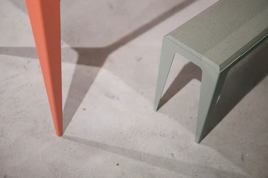Nahaufnahme von zwei modernen Tischbeinen, eines orange und das andere grün, auf einem hellen Untergrund, mit langen Schatten, die den minimalistischen Stil unterstützen. Das Design könnte zur "wye not"-Kampagne für nachhaltige Möbel passen.