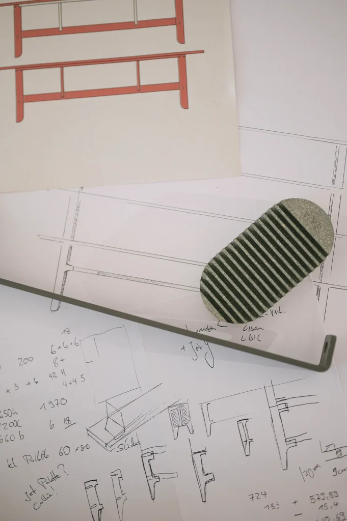 Auf einem Tisch liegen technische Zeichnungen von Möbelteilen, darunter ein gezeichneter Regalentwurf in Rot. Ein Metallstück und eine Struktur mit Streifenmuster sind ebenfalls sichtbar.