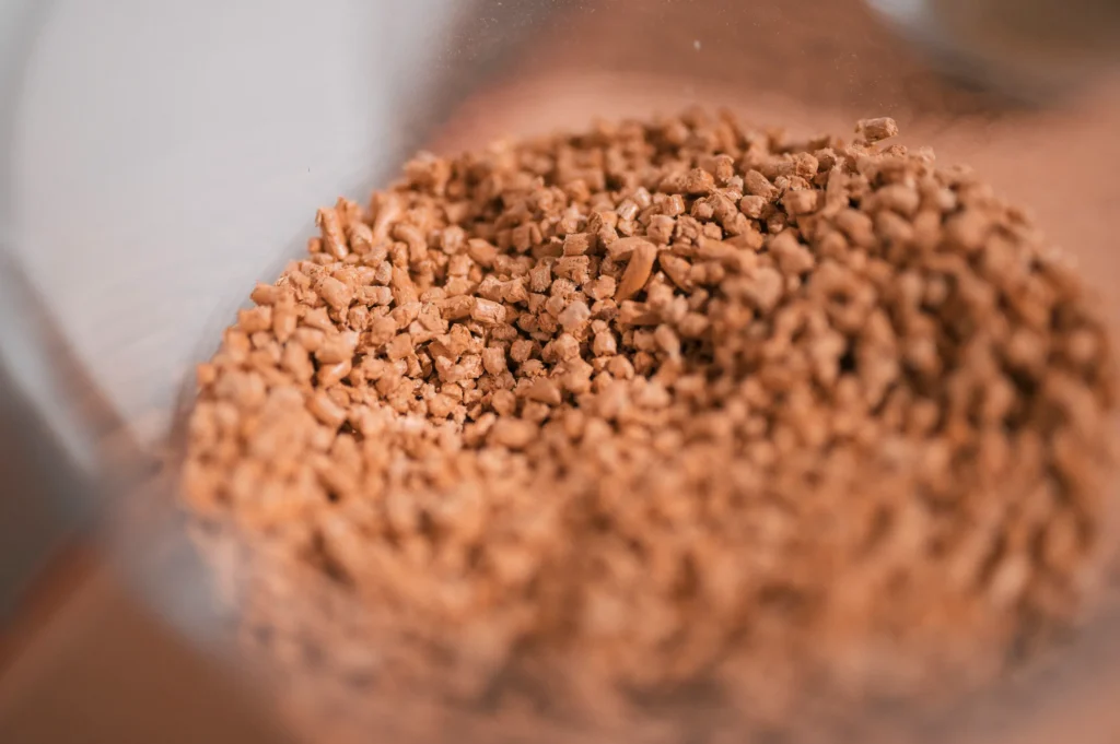 Eine Nahaufnahme von braunen, granulatförmigen Kaffeepulver in einer transparenten Schüssel. Das Pulver hat eine gleichmäßige Textur und ist gleichmäßig verteilt, das Hintergrundlicht sorgt für eine sanfte Weichzeichnung.