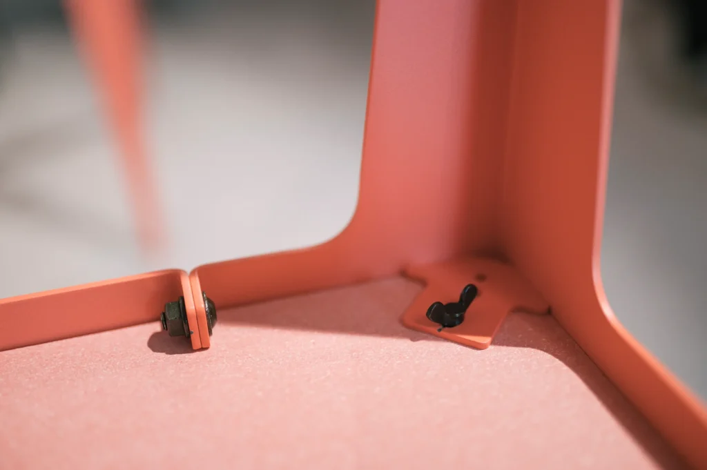 Ein Detail einer rosa farbigen Möbelkonstruktion, das die Verbindungselemente an der Basis zeigt. Es sind Schrauben und ein Schalter sichtbar, die zur Stabilität und Funktionalität beitragen. Das Design spiegelt zeitgemäße Ästhetik wider.