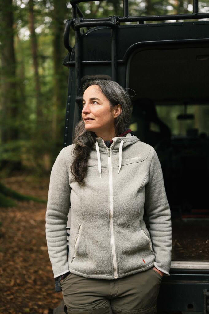 Eine Person mit langen, grauen Haaren steht neben einem Fahrzeug in einem Waldgebiet. Sie trägt eine graue Jacke und schaut nachdenklich in die Ferne. Der Boden ist mit Laub bedeckt.