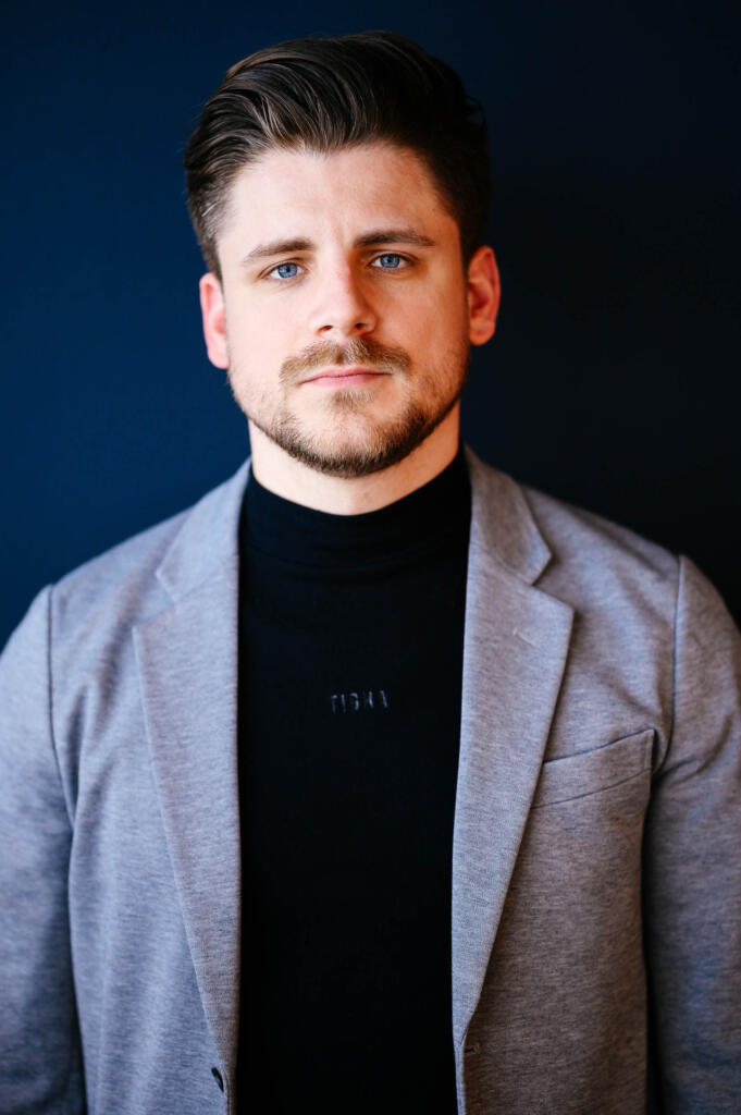 Ein Mann mit kurzen, gestylten Haaren und Bart, der in einem grauen Blazer und einem schwarzen Rollkragenpullover vor einem dunkelblauen Hintergrund steht. Er schaut direkt in die Kamera.