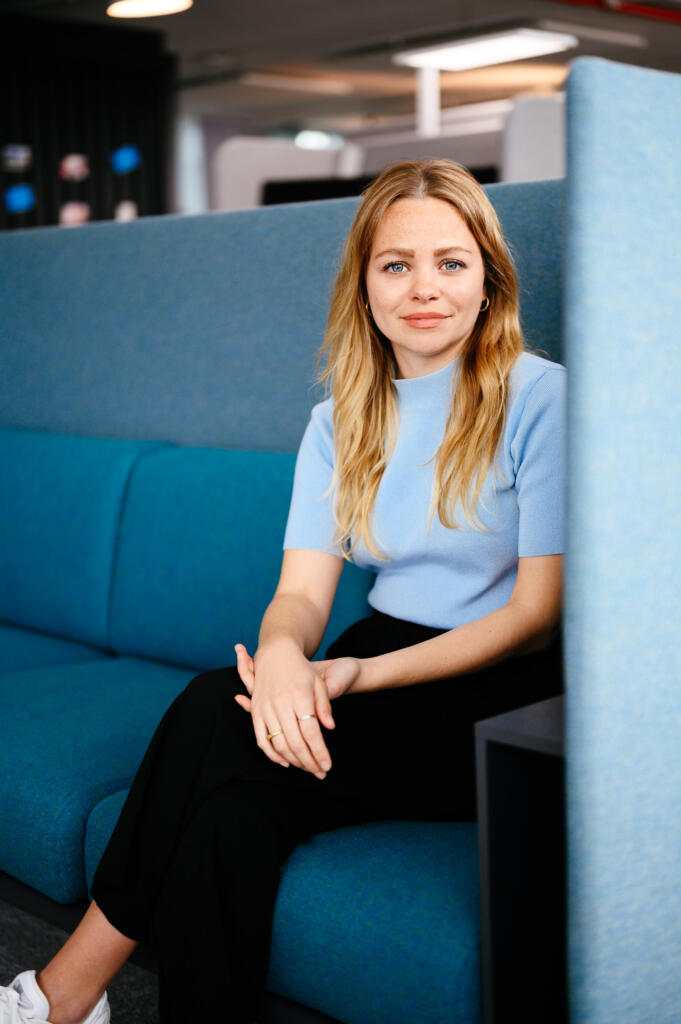 Eine Frau mit langen, blonden Haaren sitzt auf einer blauen Couch in einem modernen Büro. Sie trägt ein hellblaues Oberteil und schwarze Hosen. Im Hintergrund sind verschwommene Büromöbel sichtbar.