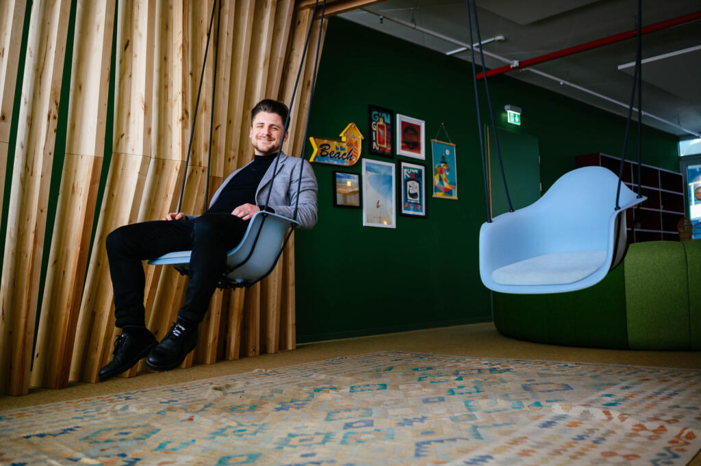 Ein Mann sitzt auf einer hängenden Sitzgelegenheit in einem modernen Büro. Im Hintergrund sind Holzvertäfelungen und verschiedene Bilder an der Wand sichtbar. Der Raum hat eine grüne Wand und einen farbenfrohen Bodenbelag.