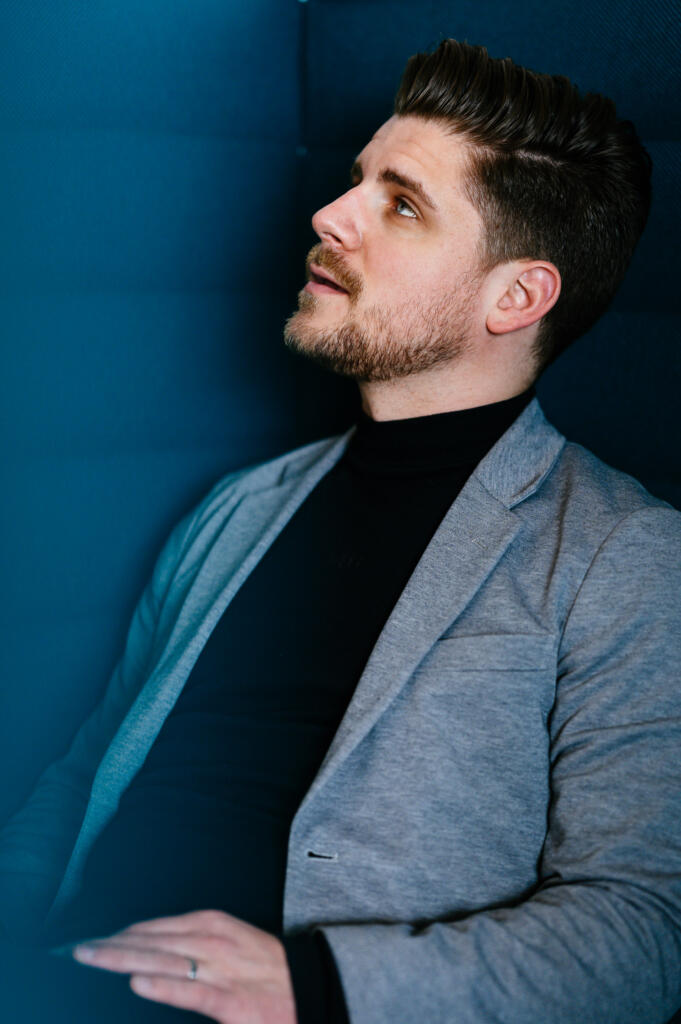 Ein Mann mit dunklem T-Shirt und grauem Blazer schaut nach oben, während er in einem modernen, blauen Raum sitzt. Seine Gesichtszüge sind konzentriert und nachdenklich.