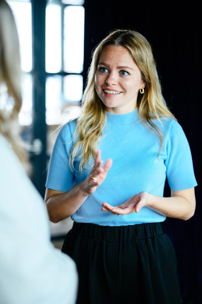 Eine lächelnde Frau mit langen, blonden Haaren trägt ein hellblaues Oberteil und einen schwarzen Rock. Sie spricht und gestikuliert in einem hellen Raum mit Fenstern im Hintergrund.
