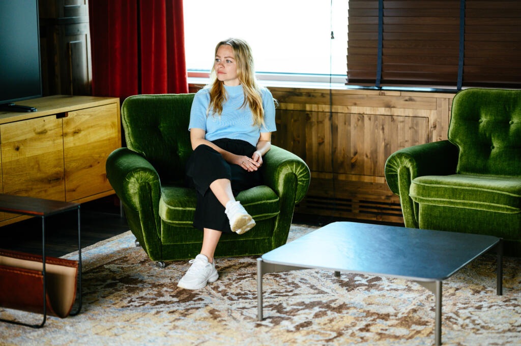 Eine Frau sitzt auf einem grünen Sessel in einem modernen Raum mit Holzmöbeln. Sie trägt ein blaues T-Shirt und schwarze Hosen. Vor ihr steht ein niedriger Tisch. Im Hintergrund sind rote Vorhänge und ein Fenster sichtbar.