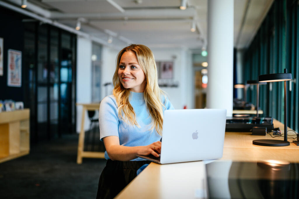 Eine Frau mit langen blonden Haaren sitzt an einem Schreibtisch mit einem Laptop in einem modernen Büro. Sie trägt ein hellblaues Oberteil und lächelt in die Kamera. Im Hintergrund sind Bücherregale und eine große Fensterfront zu sehen.