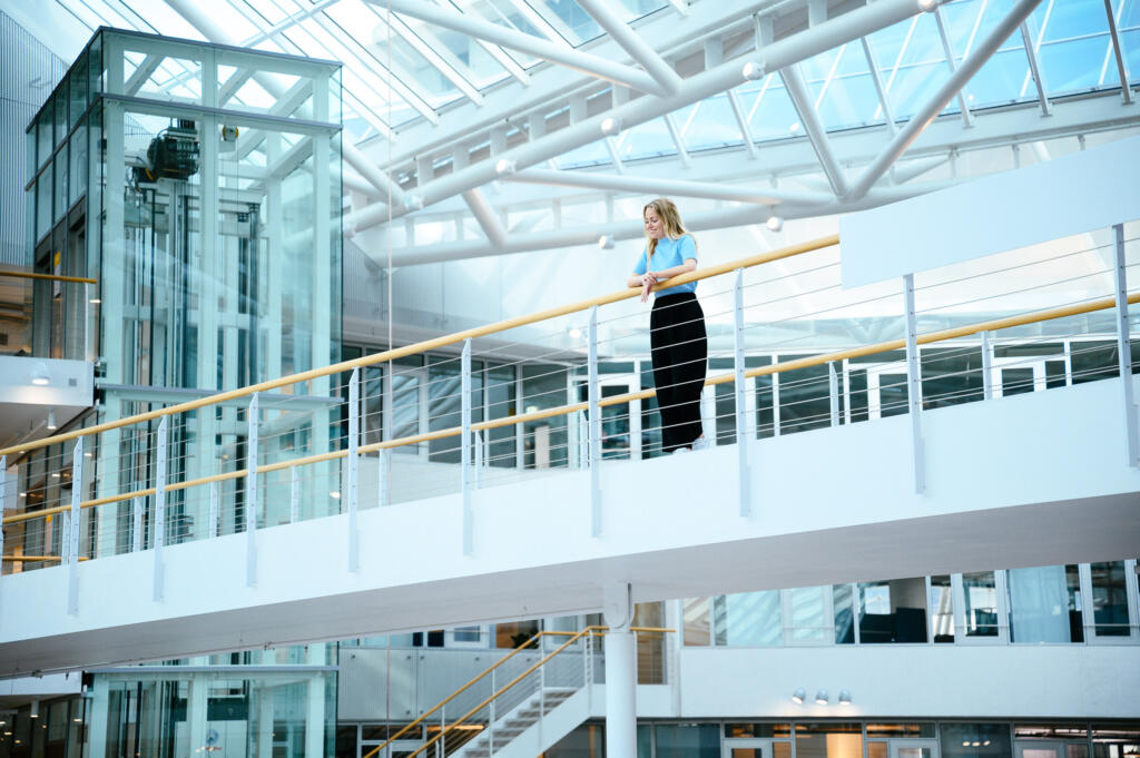 Frau in blauer Bluse und schwarzer Hose steht an einem Geländer in einem modernen Bürogebäude mit Glasarchitektur. Im Hintergrund ist ein Aufzug sichtbar.