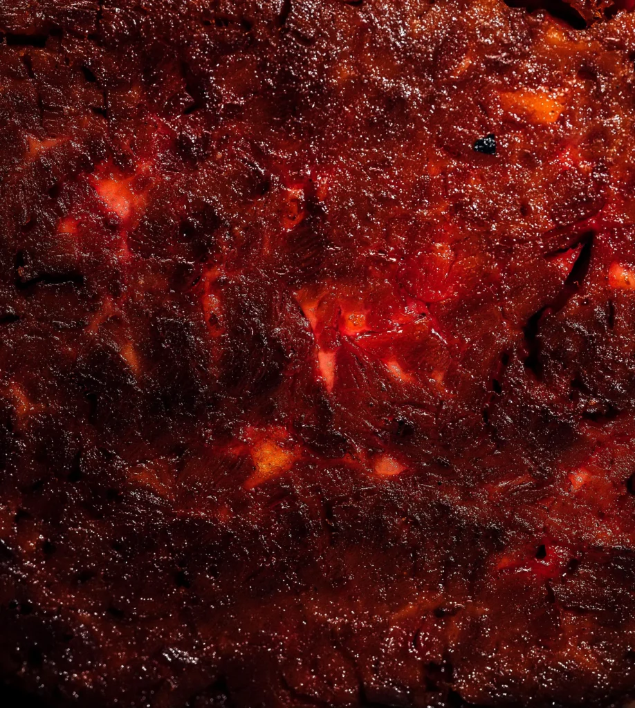 Eine oberflächennahe Aufnahme von einer dichten, dunkelbraunen Masse mit roten und orangefarbenen Stellen, die auf eine Art von Gebäck oder Konservierung hinweist.
