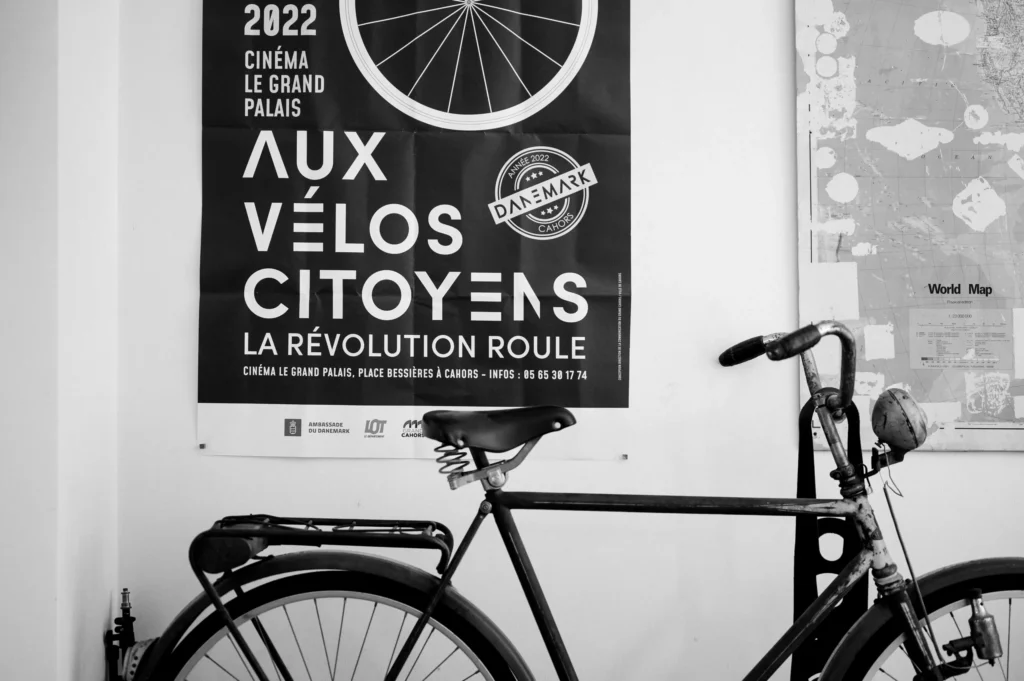 Ein schwarzes Fahrrad lehnt an einer Wand, neben einem Plakat für eine Filmvorführung mit dem Titel "Aux Vélos Citoyens". Das Plakat zeigt das Jahr 2022 und eine Fahrradgrafik.