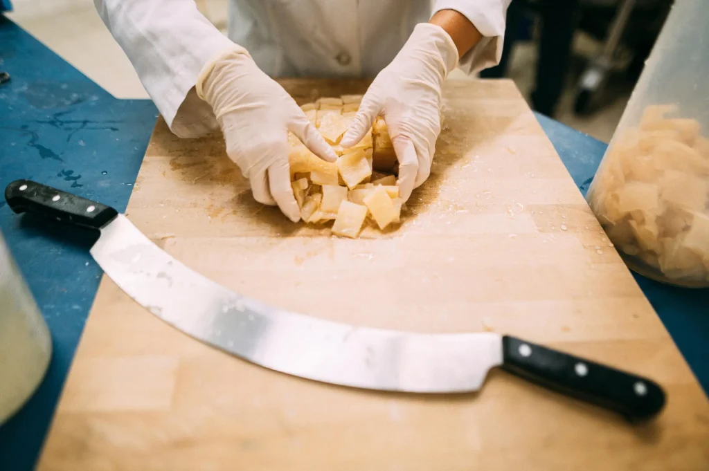 Eine Person in Arbeitshandschuhen schneidet Kartoffeln auf einem Holzbrett mit einem großen Messer. Der Hintergrund zeigt eine moderne Küchenumgebung.