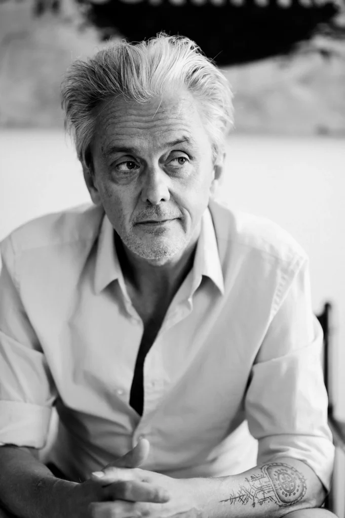 Ein Mann mit grauen Haaren sitzt an einem Tisch. Er trägt ein offenes, helles Hemd und hat seine Hände gefaltet. Der Hintergrund zeigt unscharfe, abstrakte Elemente.