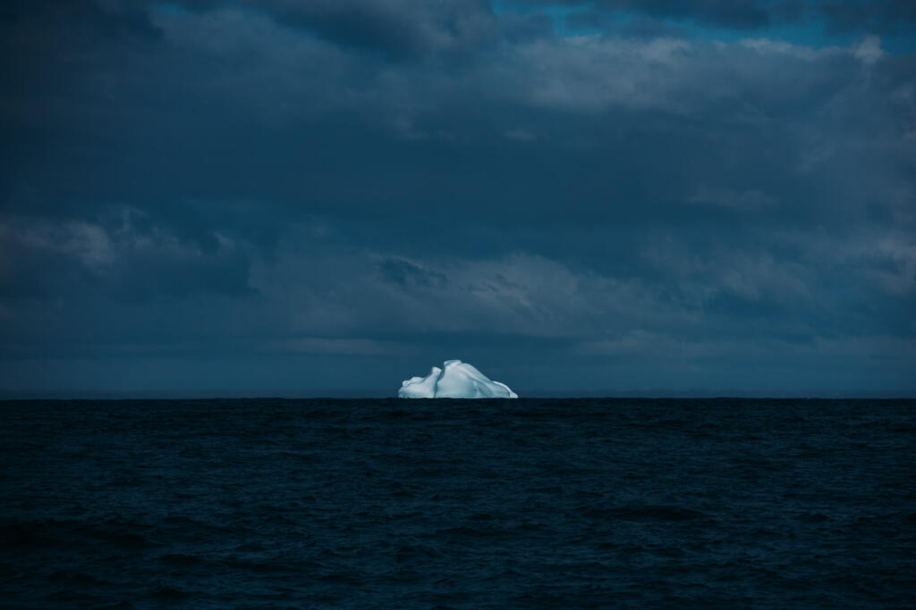 Ein einzelner Eisberg, der aus dem Ozean ragt, unter einem bewölkten, dunklen Himmel. Die Wasseroberfläche ist ruhig und reflektiert das schwache Licht.