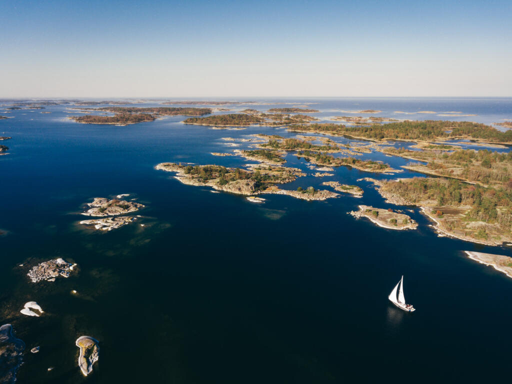 Luftaufnahme eines klaren Gewässers mit vielen kleinen Inseln und einer Segeljacht, die ruhig auf dem Wasser segelt. Der Himmel ist blau und wolkenlos.