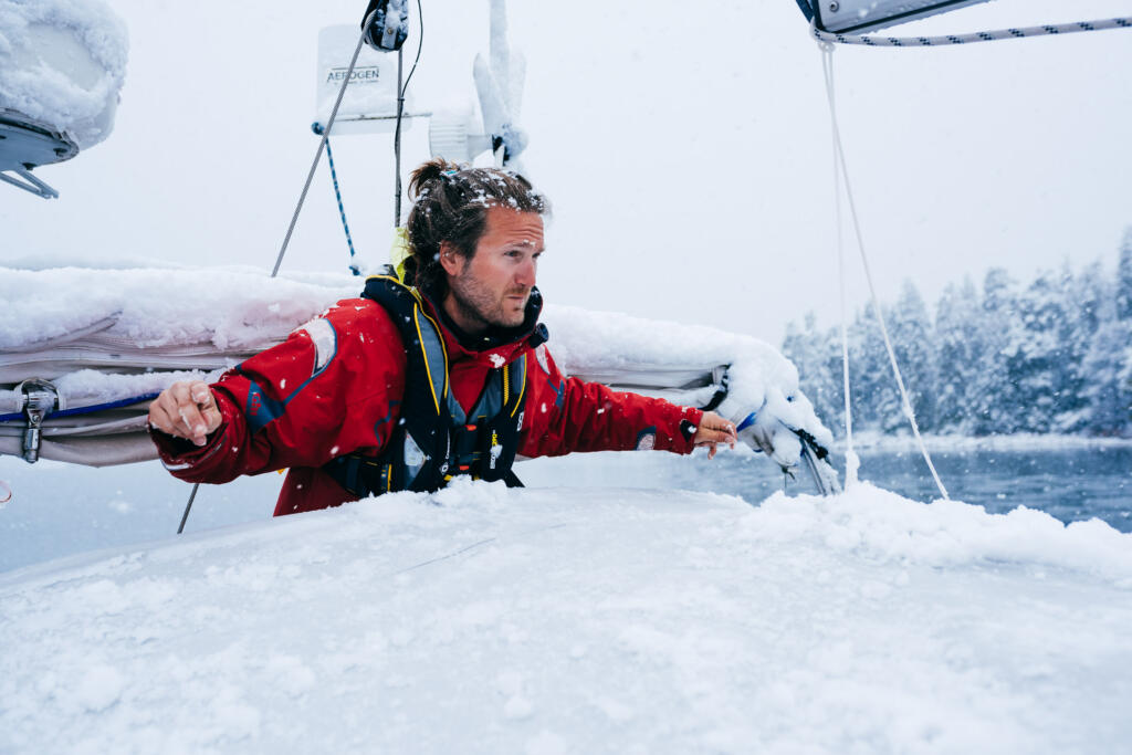 Ein Mann in roter Winterbekleidung steht auf einem segelboot, das mit Schnee bedeckt ist. Er schaut ernsthaft auf das Wasser, während es schneit und die umliegende Landschaft bewaldet ist.