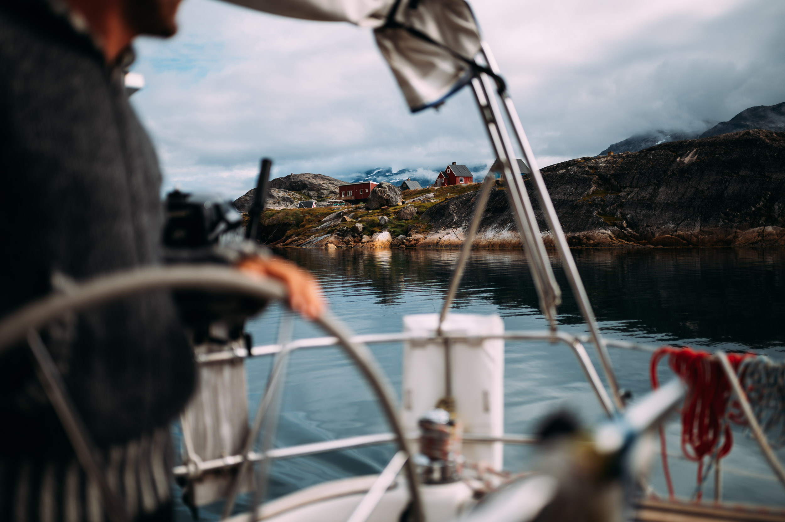 Ein Mann, der das Ruder eines Bootes hält, während er eine ruhige Wasseroberfläche mit einer Felsenküste im Hintergrund durchfährt. Im Hintergrund sind Häuser und Berge bei bewölktem Himmel zu sehen.