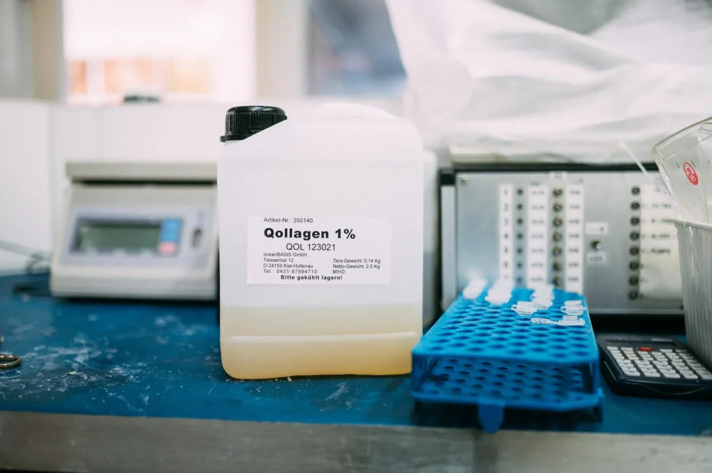 Ein Behälter mit 1% Kollagen auf einem Tisch in einem Labor. Im Hintergrund sind Laborgeräte und ein Ablesegerät sichtbar. Der Behälter hat ein Etikett mit Informationen zum Inhalt.