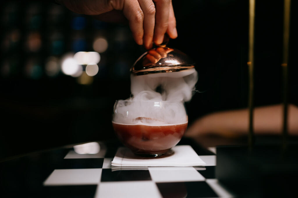 Eine Hand hebt einen Deckel von einem Glas mit rauchigem roten Getränk. Das Glas steht auf einem schwarz-weißen Tisch. Im Hintergrund sind verschwommene Lichter zu sehen.