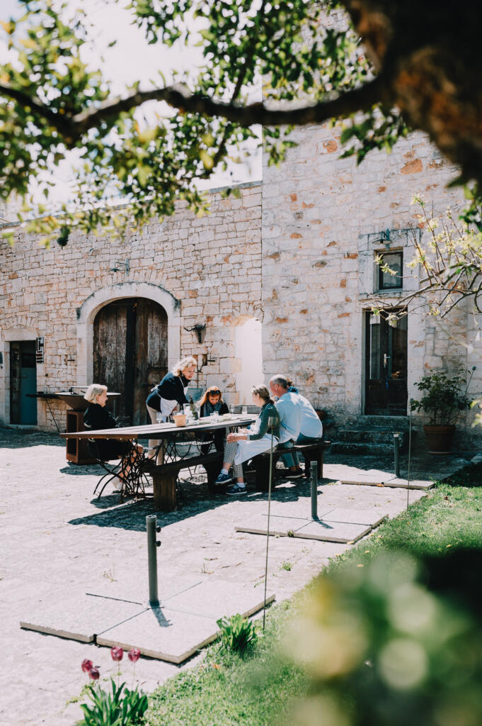 Eine Gruppe von fünf Personen sitzt an einem Tisch im Freien, während sie miteinander sprechen und essen. Im Hintergrund ist ein steinernes Gebäude mit großen Fenstern und einem Holztor zu sehen.