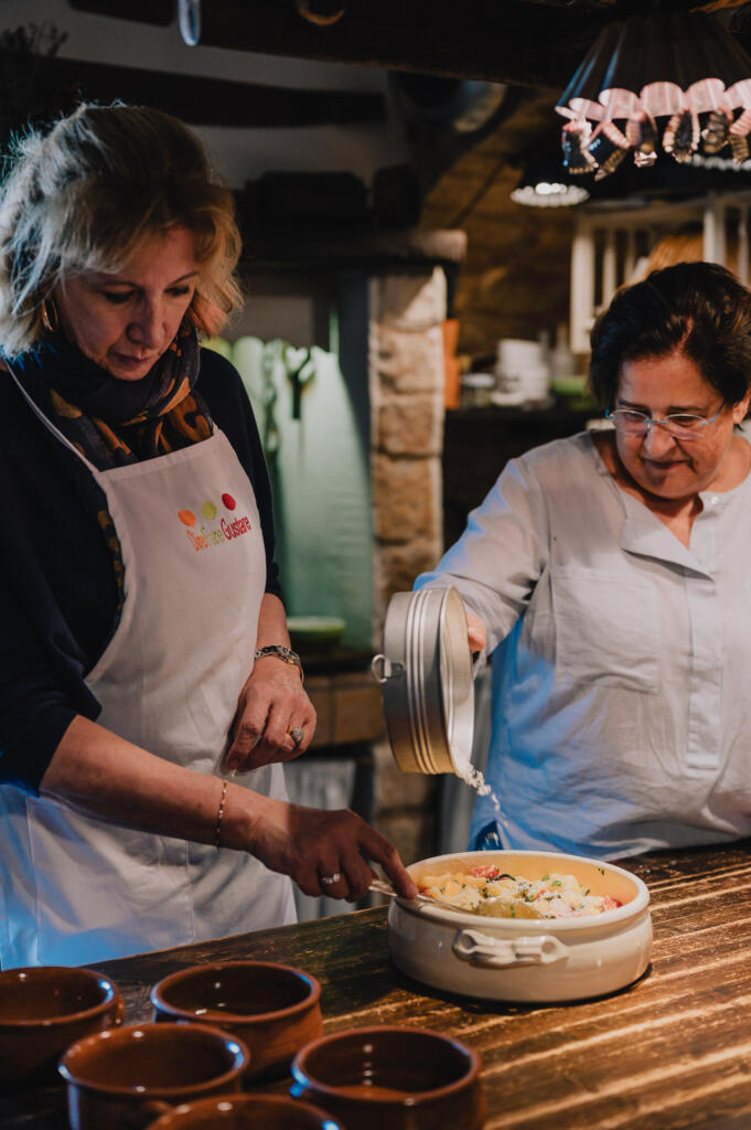Zwei Frauen in einer Küche, die an einem Kochprojekt arbeiten. Eine Frau hebt den Deckel eines weißen Kochgeschirrs, während die andere mit einem Löffel etwas herausnimmt. Dekorative Küchenumgebung ist im Hintergrund sichtbar.