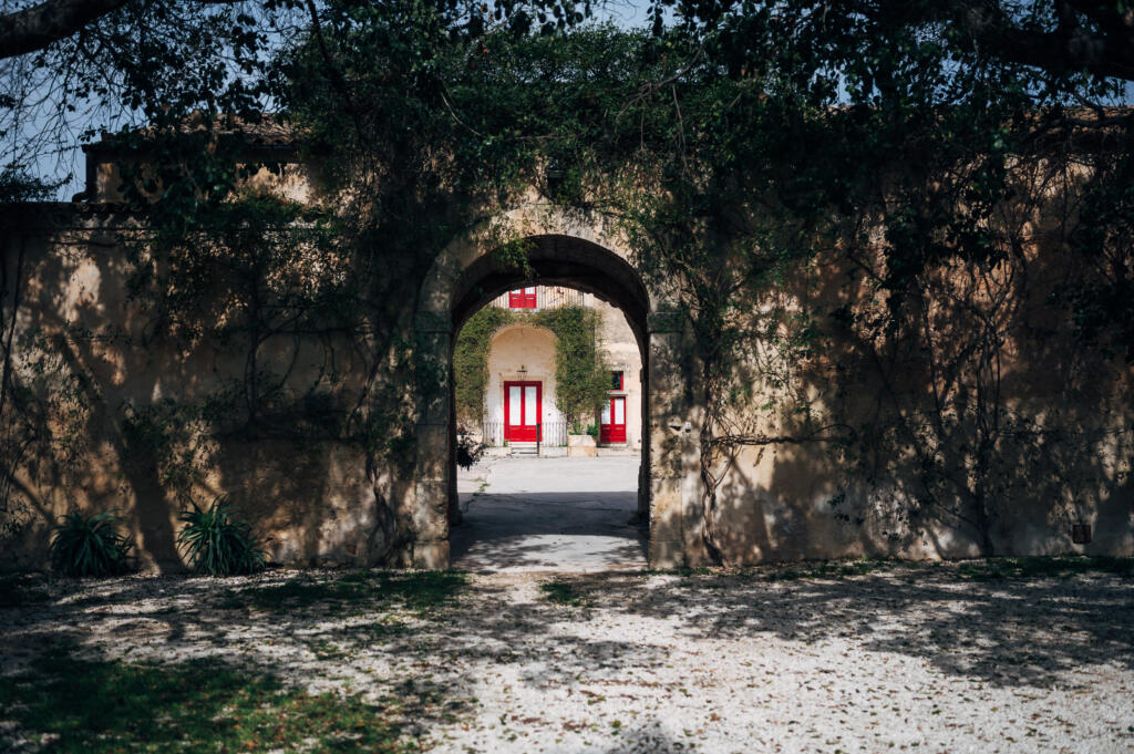 Ein Blick durch einen bogenförmigen Eingang, umgeben von grünem Efeu, auf eine gepflasterte Auffahrt mit roten Türen im Hintergrund. Der Boden ist mit Kies und Schatten von Bäumen bedeckt.