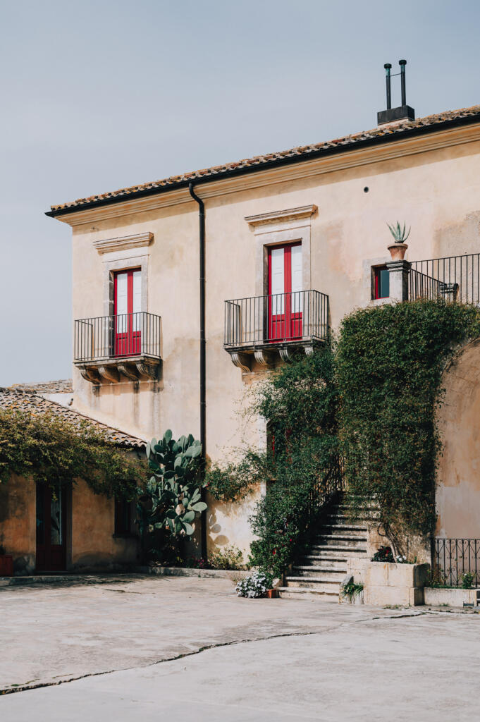 Eine mediterrane Villa mit rotem Fensterrahmen und balustradierten Balkonen, umgeben von Pflanzen und Kakteen. Ein Treppenaufgang führt zu einem Eingang, während die Fassade der Villa und der Innenhof sonnig und freundlich wirken.