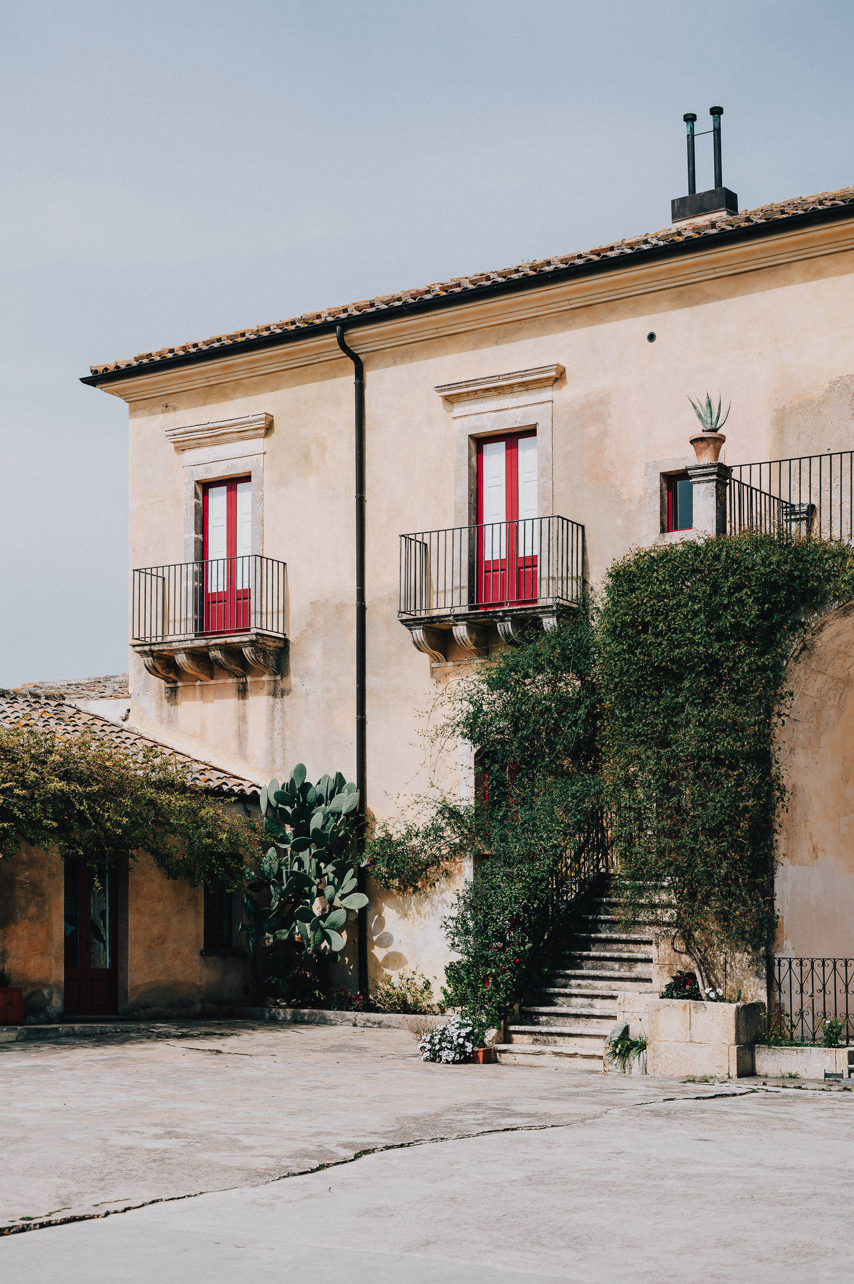 Eine mediterrane Villa mit rotem Fensterrahmen und balustradierten Balkonen, umgeben von Pflanzen und Kakteen. Ein Treppenaufgang führt zu einem Eingang, während die Fassade der Villa und der Innenhof sonnig und freundlich wirken.