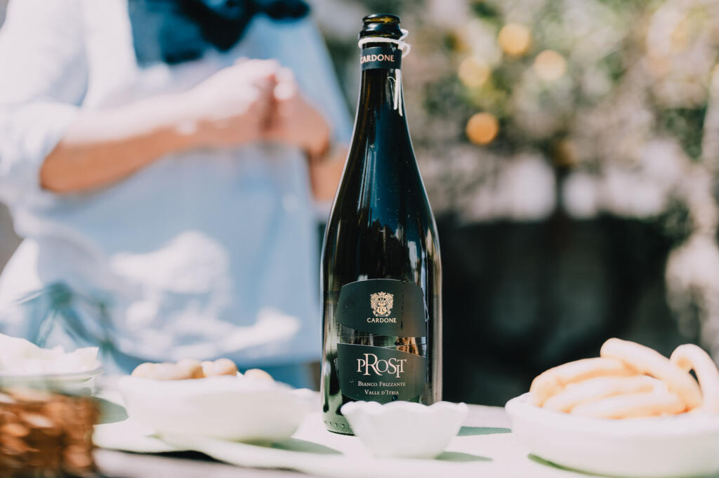 Eine Flasche Sekt mit dem Etikett "Prost" steht auf einem Tisch. Im Hintergrund sind unscharf Snacks und eine Person mit verschränkten Händen zu sehen.