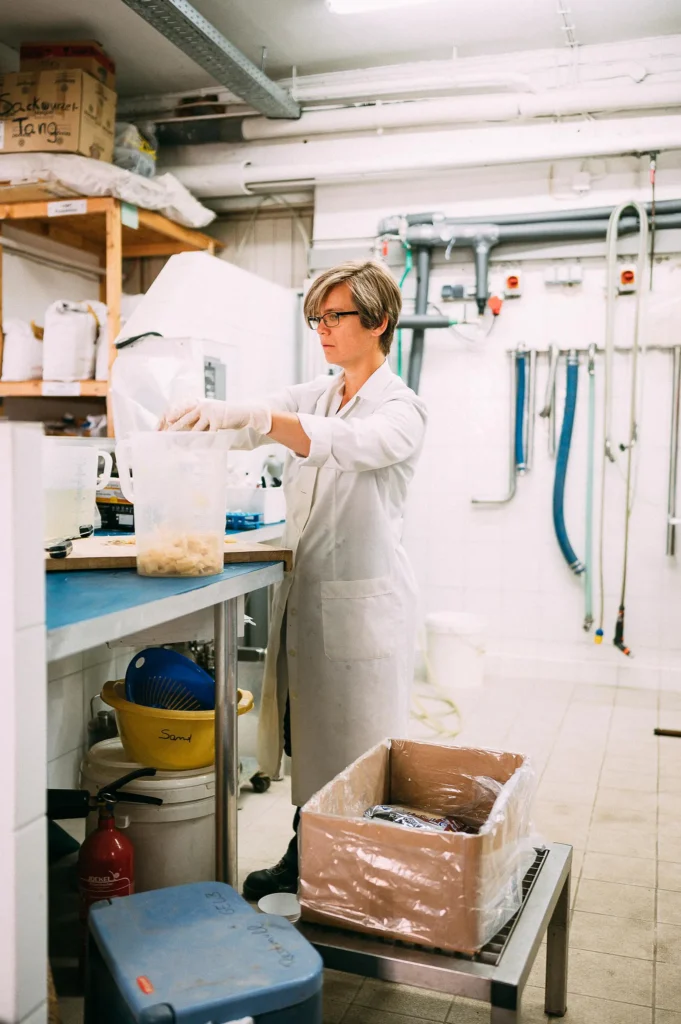 Eine Frau in einem weißen Laborkittel untersucht Proben in einem Labor mit verschiedenen Geräten und Utensilien auf einem Tisch.