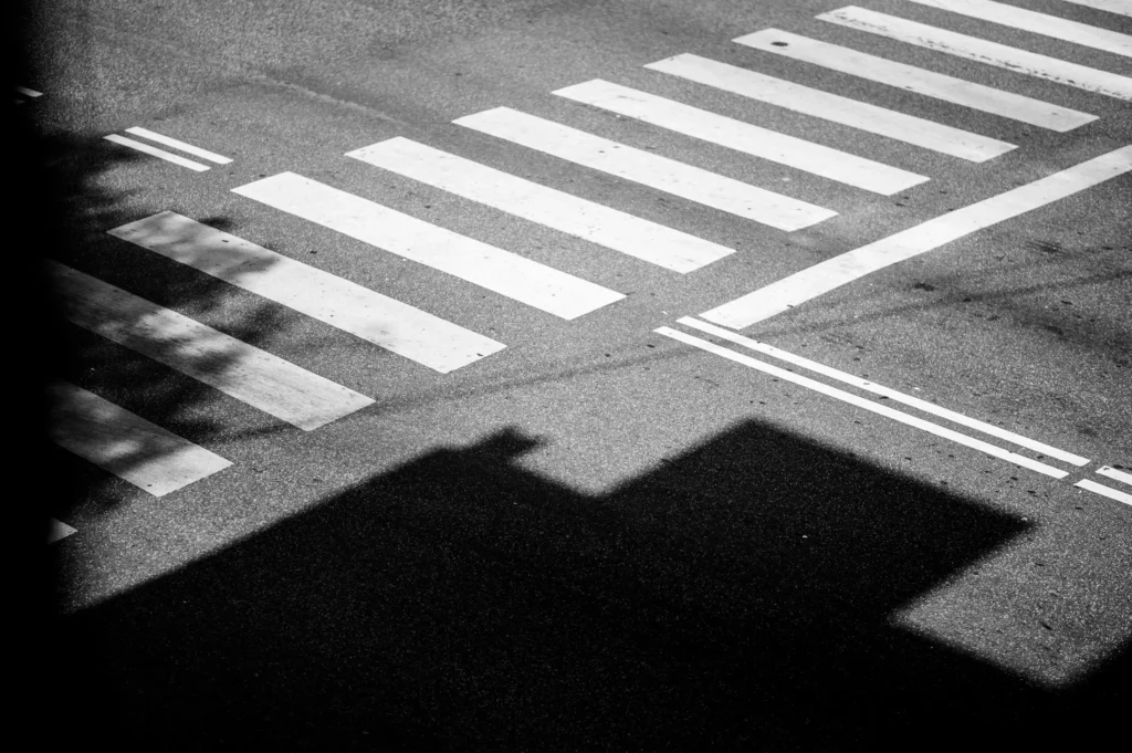 Eine Vogelperspektive auf einen Zebrastreifen mit weißen Streifen auf einer asphaltierten Straße. Im Hintergrund sind Schatten eines Gebäudes und Bäume sichtbar.