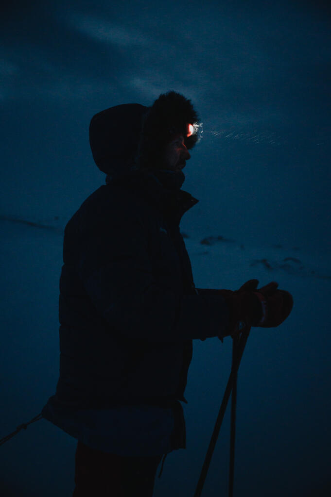 Eine Person mit einer Stirnlampe, die in der Dämmerung auf einem verschneiten Gelände steht. Die Silhouette der Person ist gegen den dunklen Himmel sichtbar, während die Umgebung schwach beleuchtet ist.