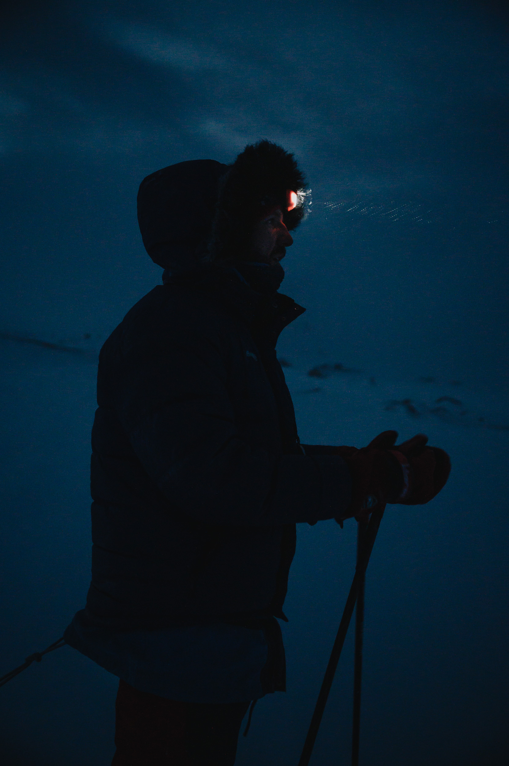 Eine Person mit einer Stirnlampe, die in der Dämmerung auf einem verschneiten Gelände steht. Die Silhouette der Person ist gegen den dunklen Himmel sichtbar, während die Umgebung schwach beleuchtet ist.