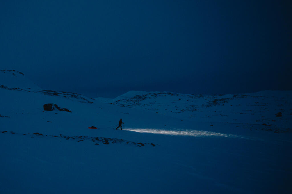 Eine Person mit einer Taschenlampe in einer schneebedeckten Landschaft bei Dunkelheit, umgeben von Bergen unter einem dunklen Himmel. Es ist eine ruhige Nacht in Finse.