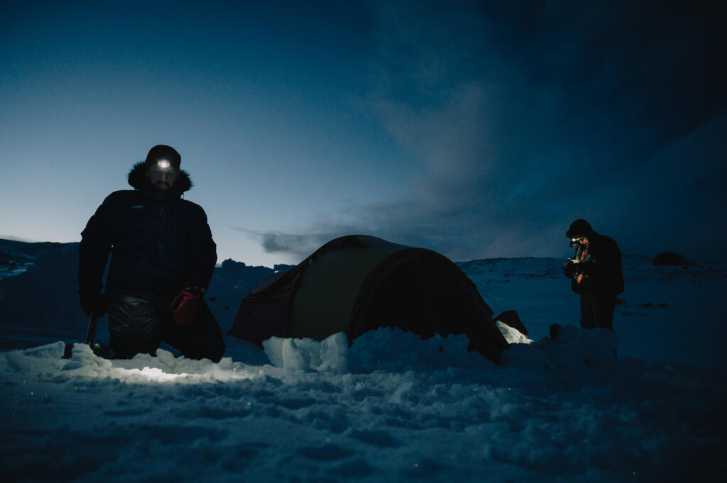 Zwei Personen im Schnee unter einem dunklen Himmel, eine Person kniet neben einem Zelt, während die andere mit einer Lampe arbeitet. Es handelt sich um eine winterliche Campingszene, möglicherweise in der Nähe von Finse.
