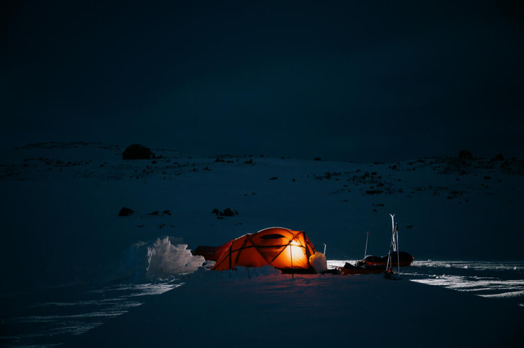 Ein orangefarbenes Zelt leuchtet in der Dunkelheit auf einem verschneiten Gelände. Um das Zelt liegt frischer Schnee, während die Umgebung karg und düster wirkt. Die Szene vermittelt eine ruhige, einsame Atmosphäre.