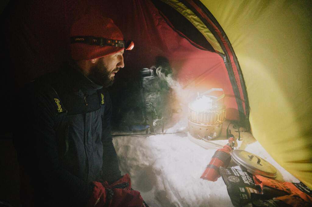 Ein Mann mit Stirnlampe in einem Zelt auf Schnee, schaut nachdenklich auf eine Wärmequelle, während dampfende Luft im Zelt sichtbar ist.