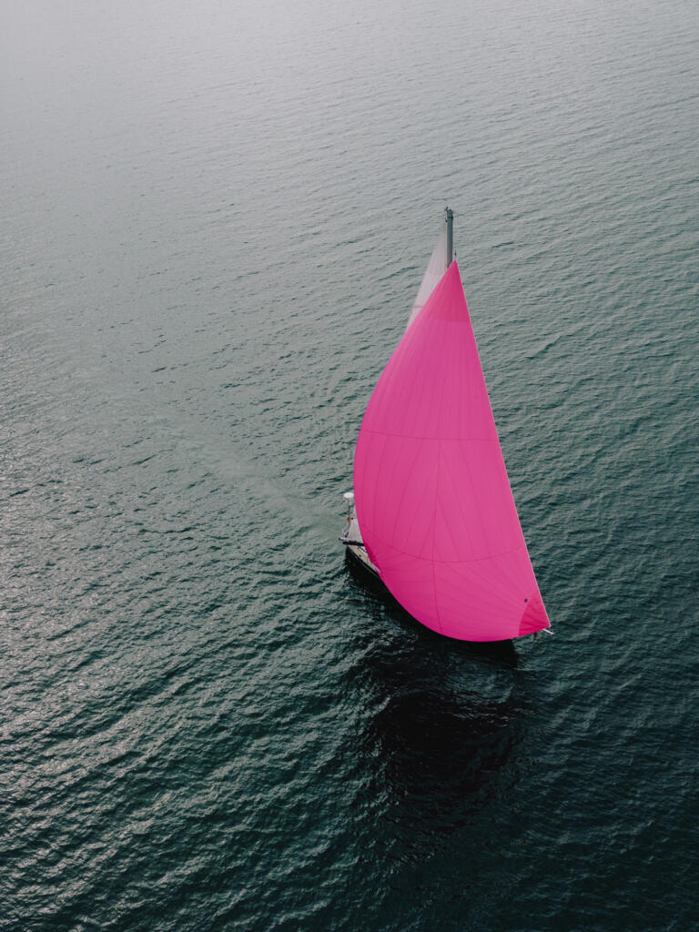 Ein segelboot mit einem großen rosa Segel fährt über das ruhige Wasser. Die Perspektive zeigt eine Luftaufnahme des Bootes und des Wassers dahinter.