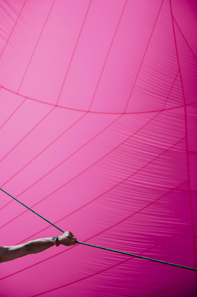 Ein naher Blick auf einen Arm, der ein Seil hält, vor einem großen, leuchtend pinkfarbenen Stoff. Die Struktur des Stoffes zeigt Falten und Linien, die durch das Licht sichtbar werden.