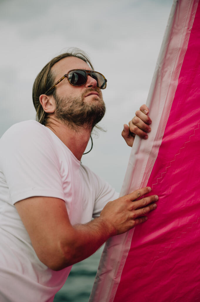 Ein Mann in Sonnenbrille hält sich an einem pinken Segel auf einem Segelboot fest, während er in den Himmel blickt. Der Hintergrund zeigt graue Wolken und Wasser.