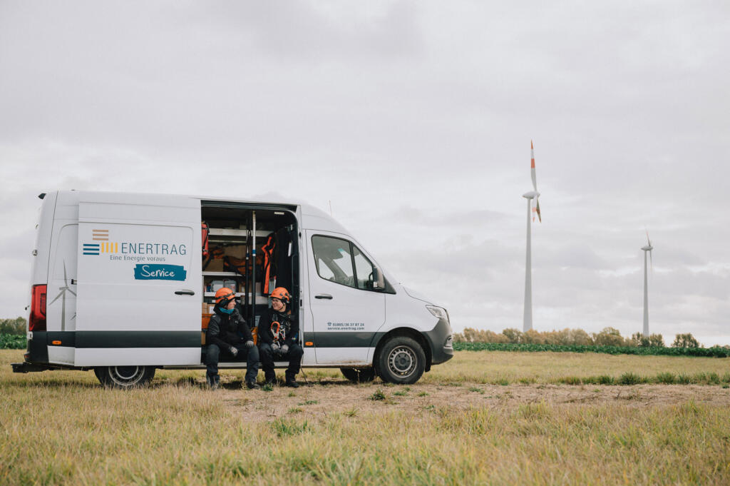 Ein Enertrag-Servicefahrzeug auf einem Feld mit zwei Arbeitern, die in Schutzkleidung sitzen. Im Hintergrund sind Windkraftanlagen sichtbar, unter einem bewölkten Himmel.