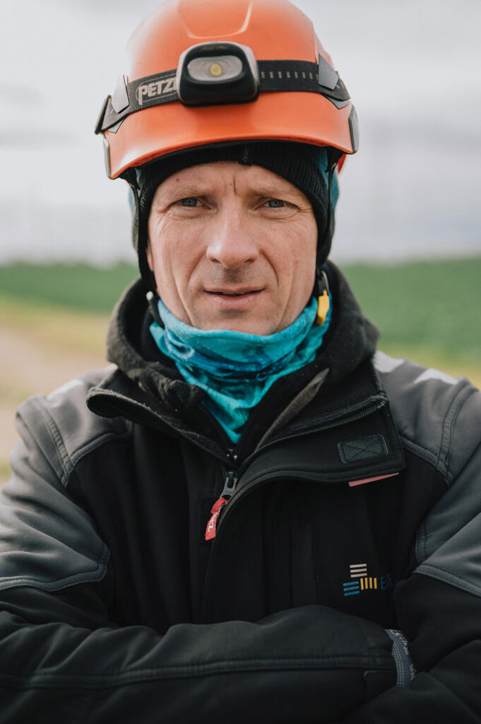 Ein Mann mit einem orangefarbenen Helm und einem blauen Schal steht mit verschränkten Armen in einer Landschaft. Er trägt eine dunkle Jacke und schaut direkt in die Kamera.
