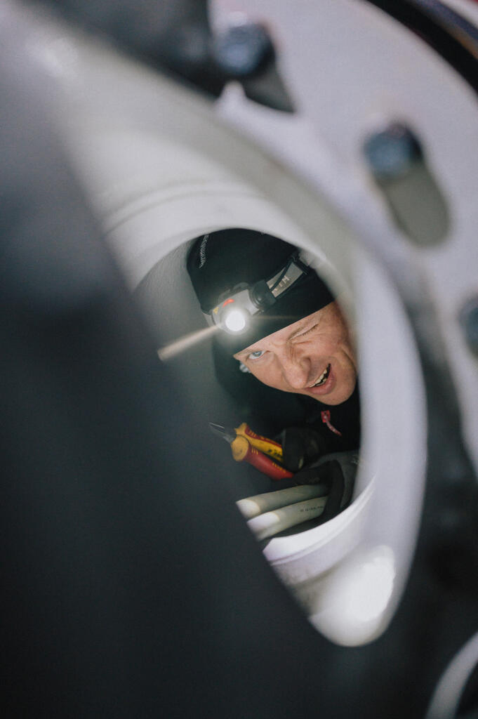 Ein Techniker mit einer Kopflampe schaut durch eine Öffnung in einer Maschine. Sein Gesicht ist teilweise sichtbar und er zeigt ein Augenzwinkern, während er mit Werkzeug in der Hand arbeitet. Das Bild repräsentiert das Engagement in der Technik bei Enertrag und unterstützt das Employer Branding.