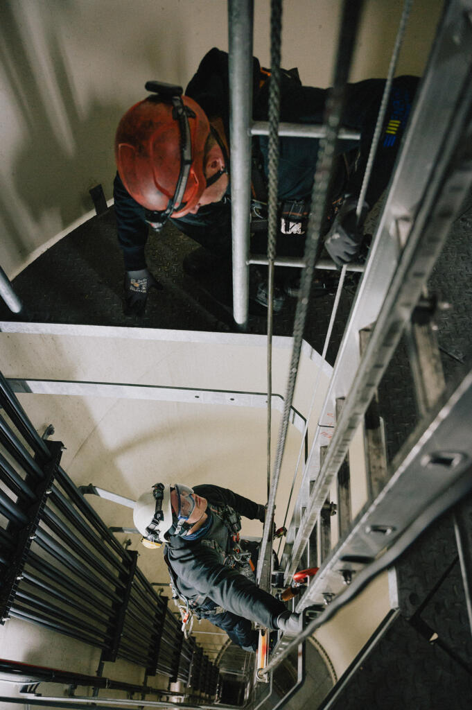 Zwei Personen arbeiten in einem engen Treppenhaus. Eine Person klettert an einer Leiter nach oben, während die andere Person im oberen Bereich der Treppe steht. Diese Szene steht im Zusammenhang mit Enertrag und Employer Branding.